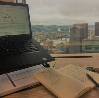 Image shows a desk with a laptop with a scientific paper opened on the screen next to a notebook and some pens. There is a window behind the desk looking out on Pittsburgh, Pennsylvania.